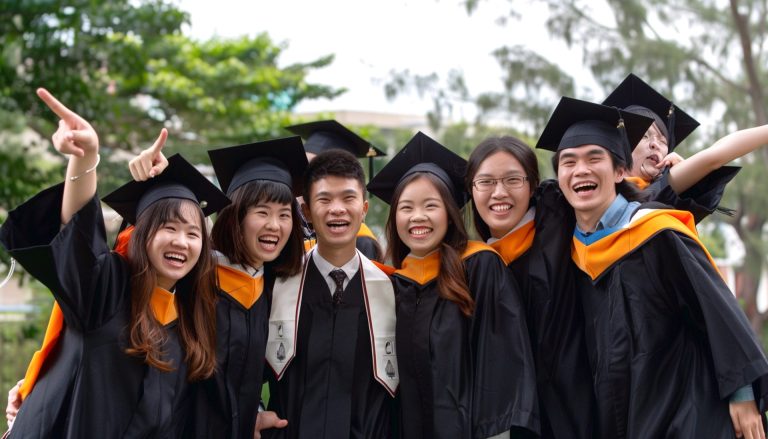 Happy grads in gowns & caps cheer outdoors together.